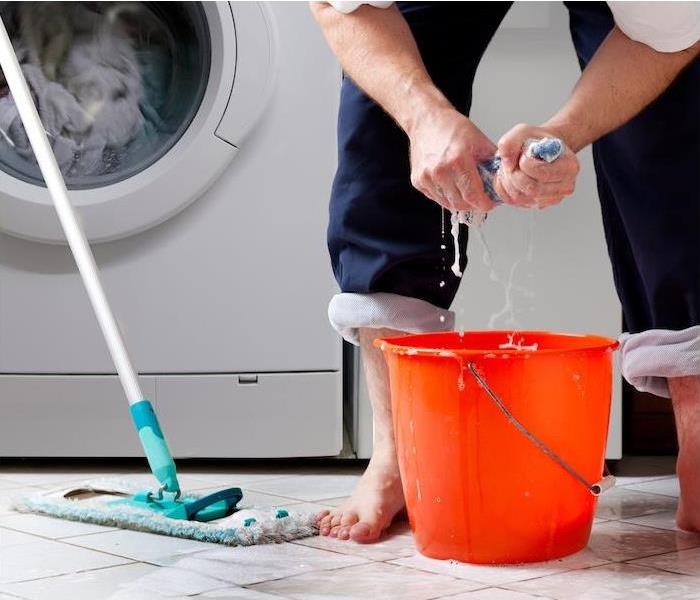 Shows a man ringing out a wet towel in a pail.  Water is covering the floor because the washing machine overflowed.